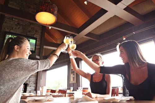 Three women clink glasses of champagne in a cozy restaurant with wooden decor and large windows.
