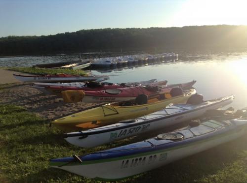 Colorful kayaks lined up on a sandy shore beside a calm lake, with trees and a dock in the background.