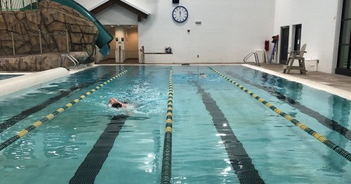 A swimming pool with lanes, featuring a water slide in the background and a clock on the wall. Two swimmers are visible.