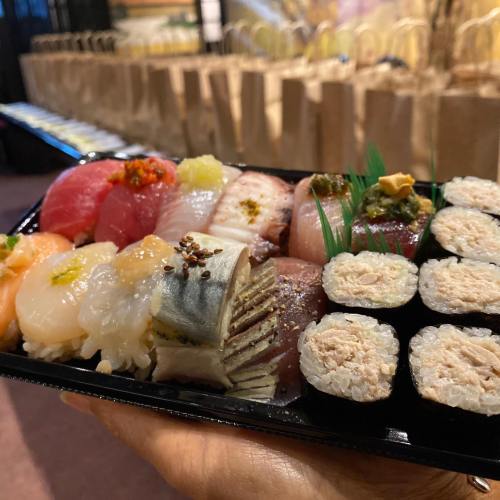 A hand holding a black sushi platter with various types of sushi and sashimi, including rolls and fresh fish.