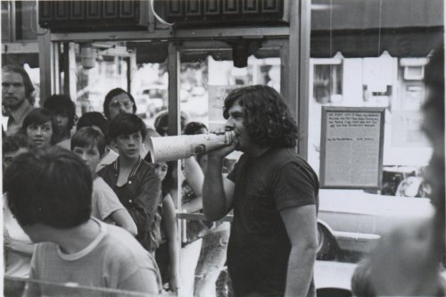 A man with long hair holds a megaphone, speaking to a crowd of onlookers outside a shop.