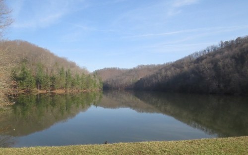 A serene lake surrounded by hills, reflecting trees and a clear blue sky.