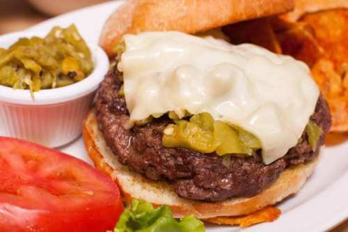 A close-up of a burger with melted cheese, green chilies, and fresh toppings, served with chips and a side of salsa.