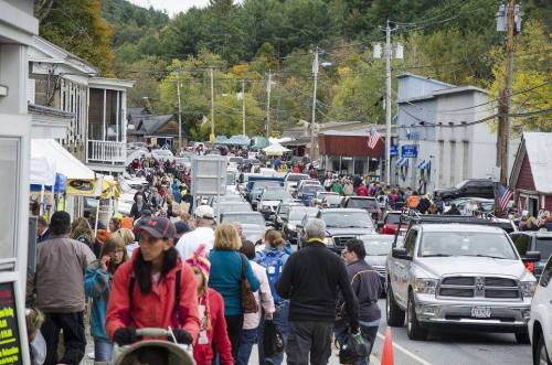 A busy street filled with people and cars, surrounded by shops and trees, during a lively outdoor event.