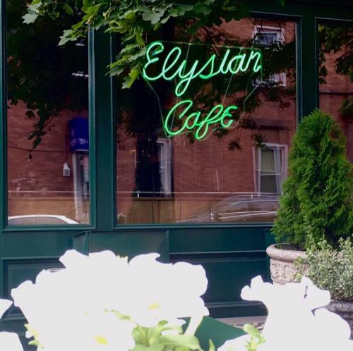Neon sign reading "Elysian Cafe" reflected in a window, surrounded by greenery and white flowers.