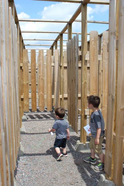 Two young boys explore a wooden maze on a sunny day, surrounded by tall wooden walls and a clear blue sky.