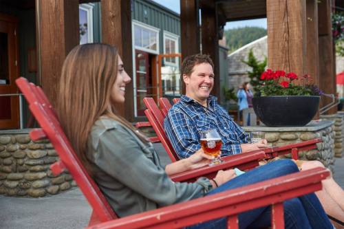 A man and woman relax in red chairs outside a building, enjoying drinks and smiling at each other. Flowers are in the background.