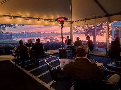A cozy tented dining area with people enjoying a meal, overlooking a sunset by the water.