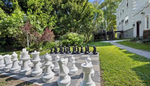 A large outdoor chess set with white and black pieces on a grassy lawn, surrounded by trees and a building in the background.