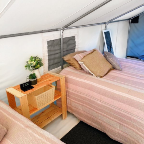 Cozy tent interior with two beds, decorative pillows, a wooden side table, and a clock.