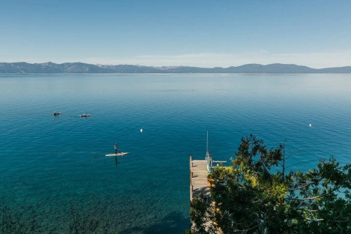 A serene lake view with calm waters, a dock, and a paddleboarder, surrounded by mountains under a clear blue sky.