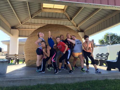 A group of women posing playfully in athletic wear in front of a large hay bale under a shelter.