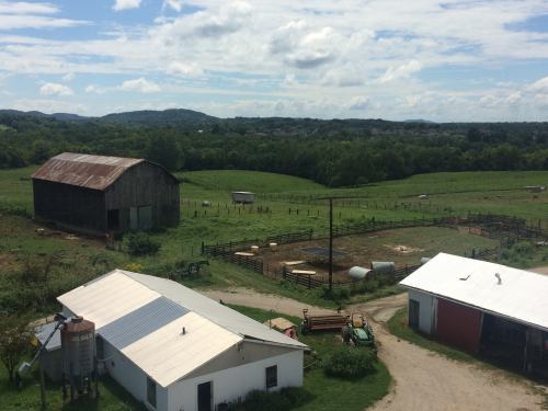 A scenic view of a farm with a barn, fields, and distant hills under a partly cloudy sky.