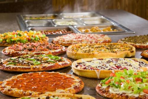 A variety of pizzas with different toppings displayed on a table, with a salad bar in the background.