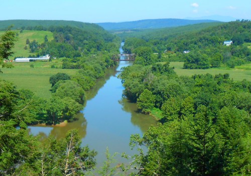 A scenic view of a winding river surrounded by lush green fields and trees, with distant hills under a clear blue sky.