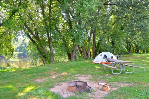 A campsite by a river featuring a tent, picnic tables, and a fire pit surrounded by lush green trees.