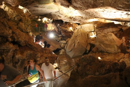 Visitors explore a rocky cave with illuminated pathways and large boulders, creating a natural underground landscape.