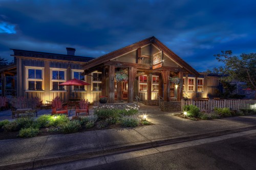 A cozy restaurant exterior at dusk, featuring wooden architecture, outdoor seating, and warm lighting.
