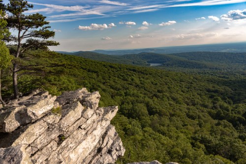 A rocky outcrop overlooks a lush green valley and distant mountains under a blue sky with scattered clouds.
