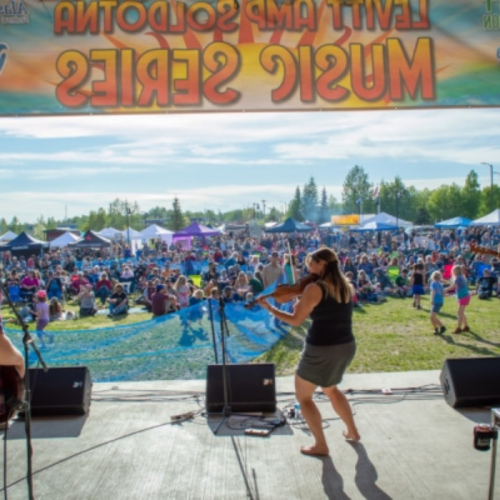 A performer plays music on stage in front of a large crowd at a lively outdoor festival. Colorful tents are visible.