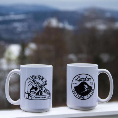 Two white mugs with logos for the Lyndon Outing Club, set against a blurred outdoor background.