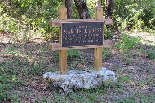 A memorial sign for Martin J. Roess, honoring his contributions to Gold Head Branch State Park, surrounded by trees.