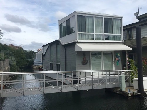 A modern houseboat with large windows, a deck, and a walkway over water, surrounded by other buildings.