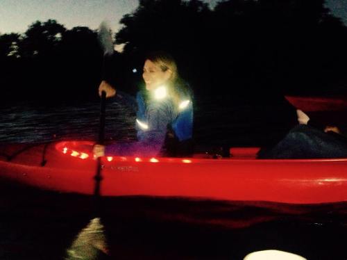 A person paddles a red kayak at dusk, wearing a reflective jacket, with lights illuminating the kayak.