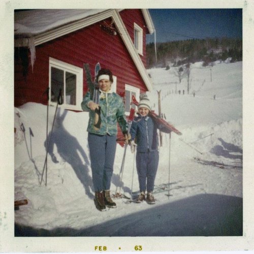 A woman and a child in winter attire jump in front of a red house, surrounded by snow and ski equipment.