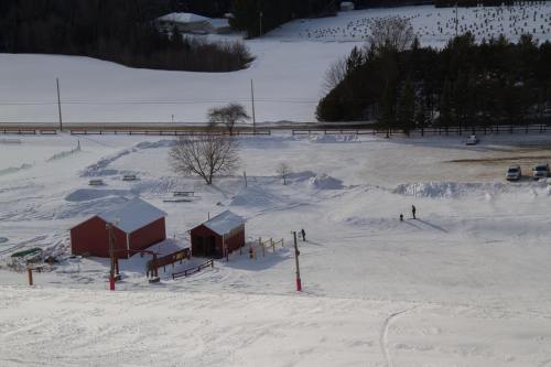 Two red cabins in a snowy landscape, with a few people walking and a tree in the background. Snow-covered fields beyond.