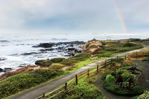 A coastal scene with rocky shores, lush greenery, a path, and a rainbow in the sky over the ocean.
