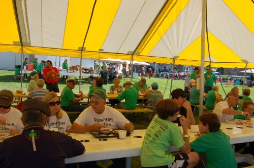 A large group of people in green shirts enjoying a meal under a yellow-striped tent at an outdoor event.