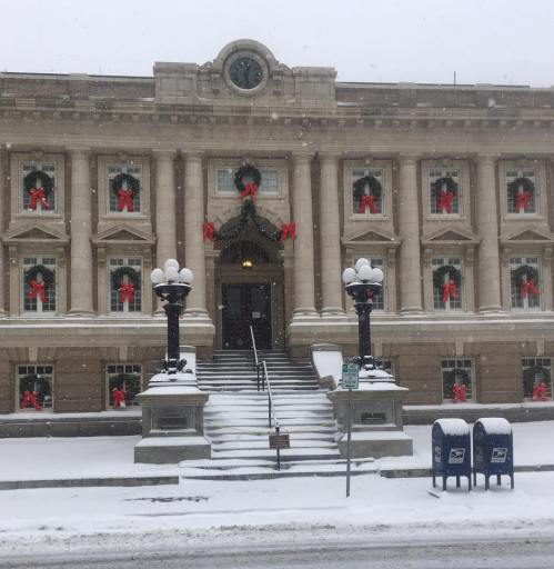 Historic building adorned with holiday wreaths, surrounded by snow, with steps leading to the entrance and a mailbox nearby.