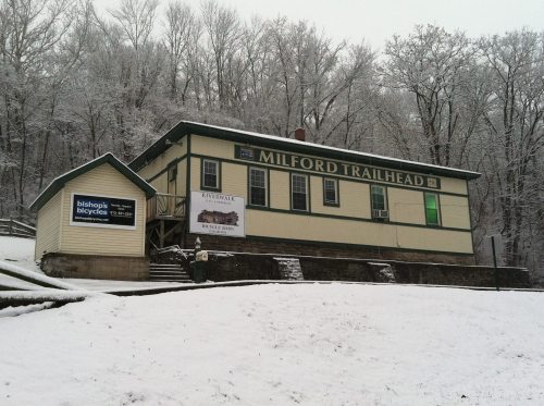 A snowy scene featuring the Milford Trailhead building with signs for bicycles and local businesses.