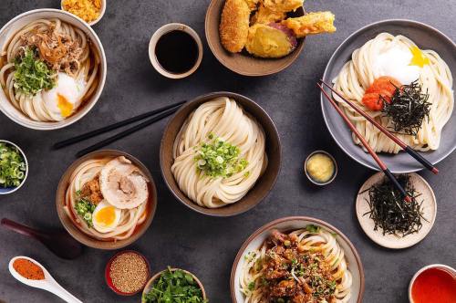A variety of noodle dishes with toppings, served in bowls, alongside small dishes of sauces and garnishes.
