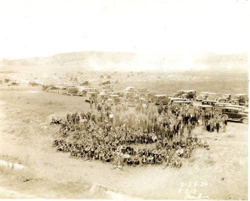 A large group of people poses for a photo outdoors, with vintage cars and a landscape in the background, dated 1936.