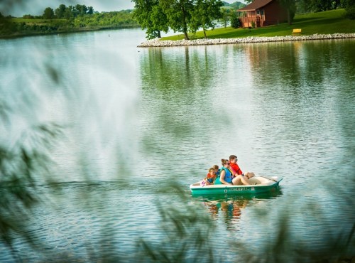 A family enjoys a boat ride on a calm lake surrounded by greenery and a rustic cabin in the background.