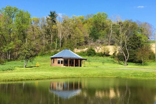 A serene landscape featuring a round cabin by a pond, surrounded by lush greenery and trees under a clear blue sky.