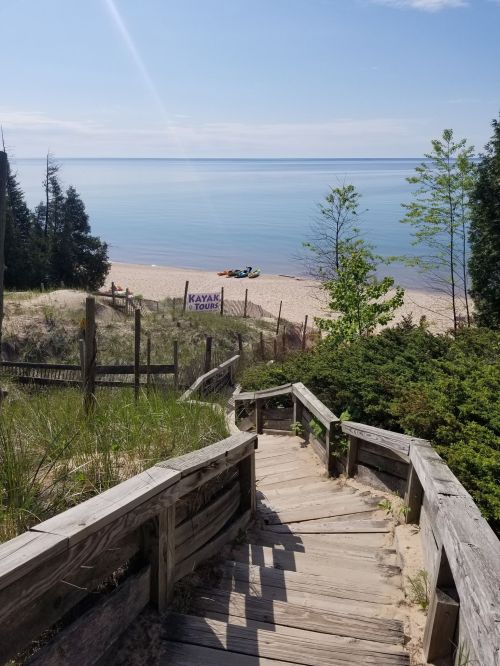 Wooden steps lead down to a sandy beach with calm water and kayaks in the distance under a clear blue sky.