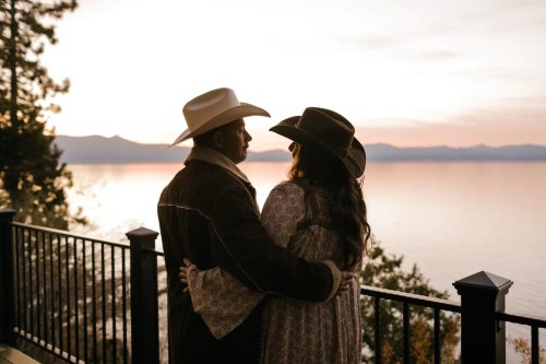 A couple in cowboy hats embraces by a lake at sunset, surrounded by trees and mountains.