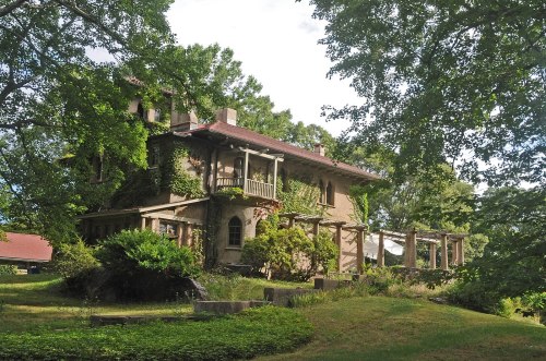 A historic stone house surrounded by lush greenery and trees, featuring a balcony and a pergola.