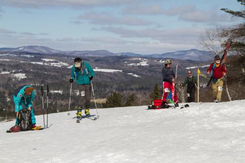 A group of skiers prepares on a snowy slope with mountains in the background under a partly cloudy sky.