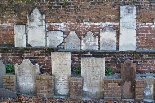 A row of old gravestones against a brick wall, some with inscriptions, surrounded by fallen leaves.