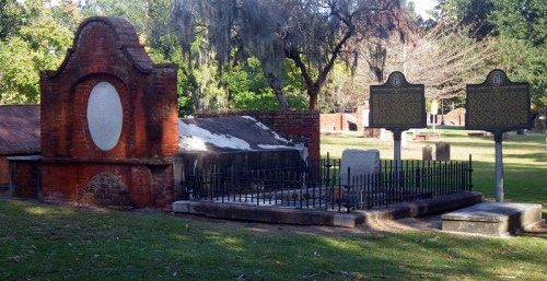 Historic cemetery scene featuring brick tombs and informational plaques amidst green grass and trees.