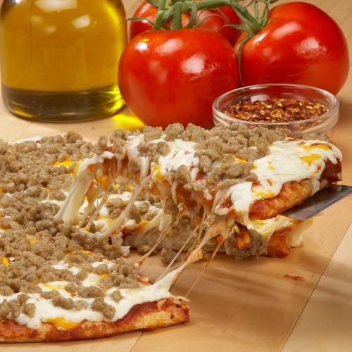 A cheesy pizza topped with sausage, pulled away to reveal gooey cheese, with fresh tomatoes and olive oil in the background.