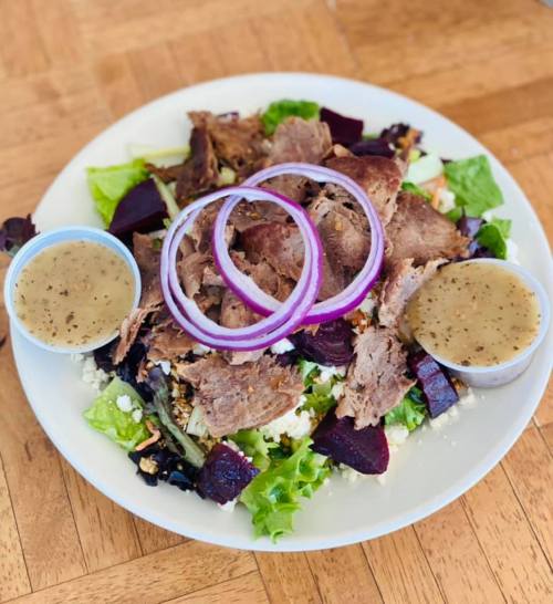 A fresh salad topped with sliced meat, red onion rings, beets, and two cups of dressing on a white plate.