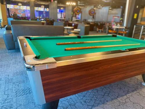A pool table with green felt and three cues, set in a lively game room with seating and dartboards in the background.