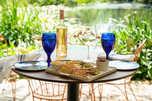 A beautifully set outdoor table with blue glasses, a bottle of rosé, and a plate of food, surrounded by greenery.