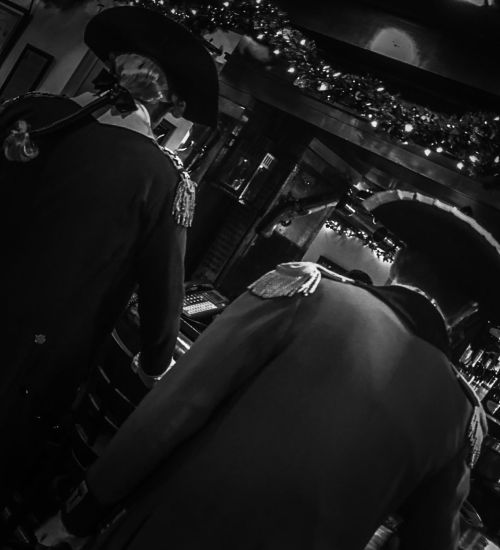 Two individuals in historical military uniforms, viewed from behind, in a dimly lit setting with festive decorations.