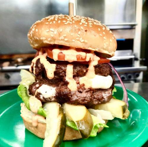 A towering burger with two patties, lettuce, tomato, pickles, and sauce, served on a sesame seed bun on a green plate.